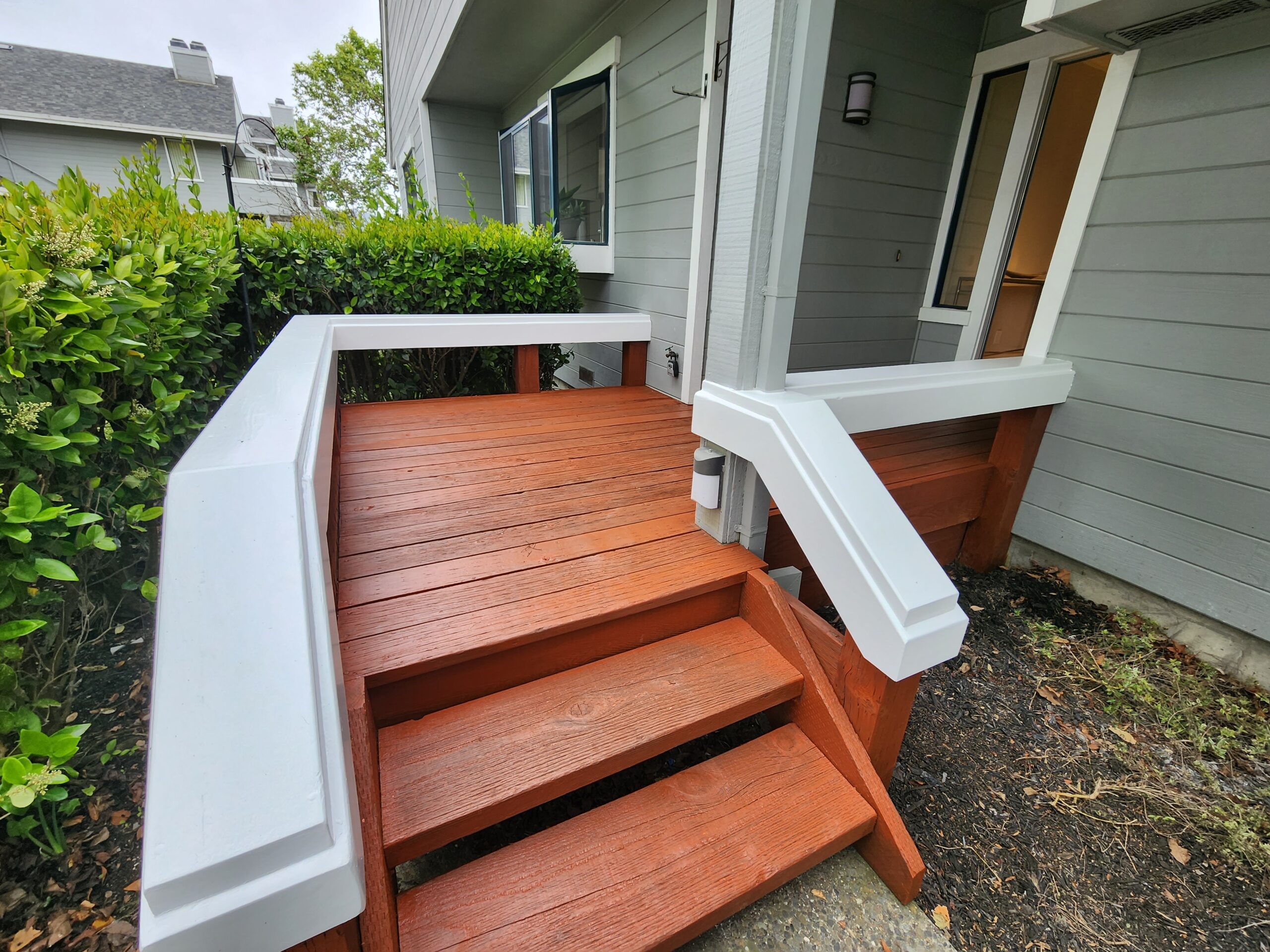A small wooden deck with red-brown stained planks and white railings leads to the entrance of a gray house with bushes on the left side.