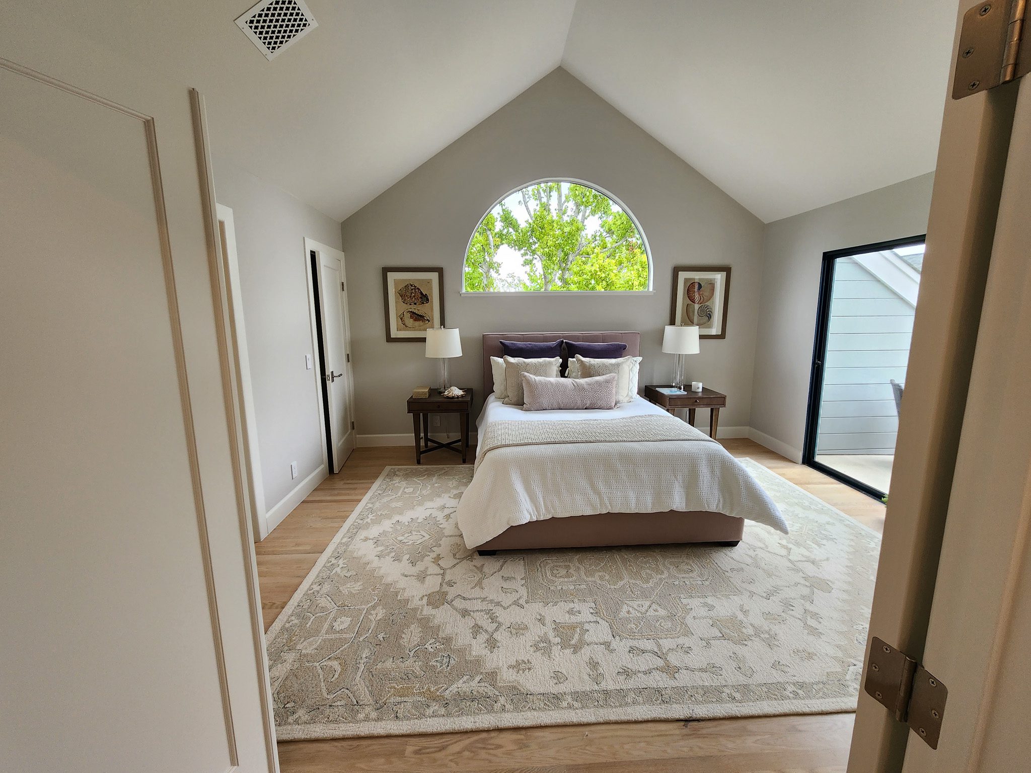 A bedroom with a slanted ceiling has a bed with white bedding, two bedside tables with lamps, a large rug, two framed pictures on the wall, and a window with a view of trees.