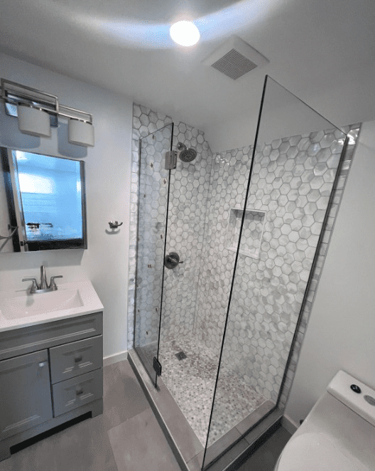 A modern bathroom featuring a glass-enclosed shower with hexagonal tile, a vanity with a mirror and light fixture, and a toilet.