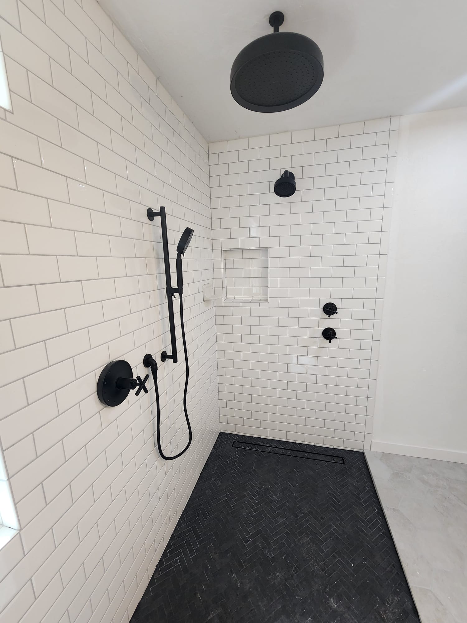 A modern shower with white subway tiles, black fixtures including a rain showerhead, hand shower, and control knobs, and a black herringbone-patterned floor mat.