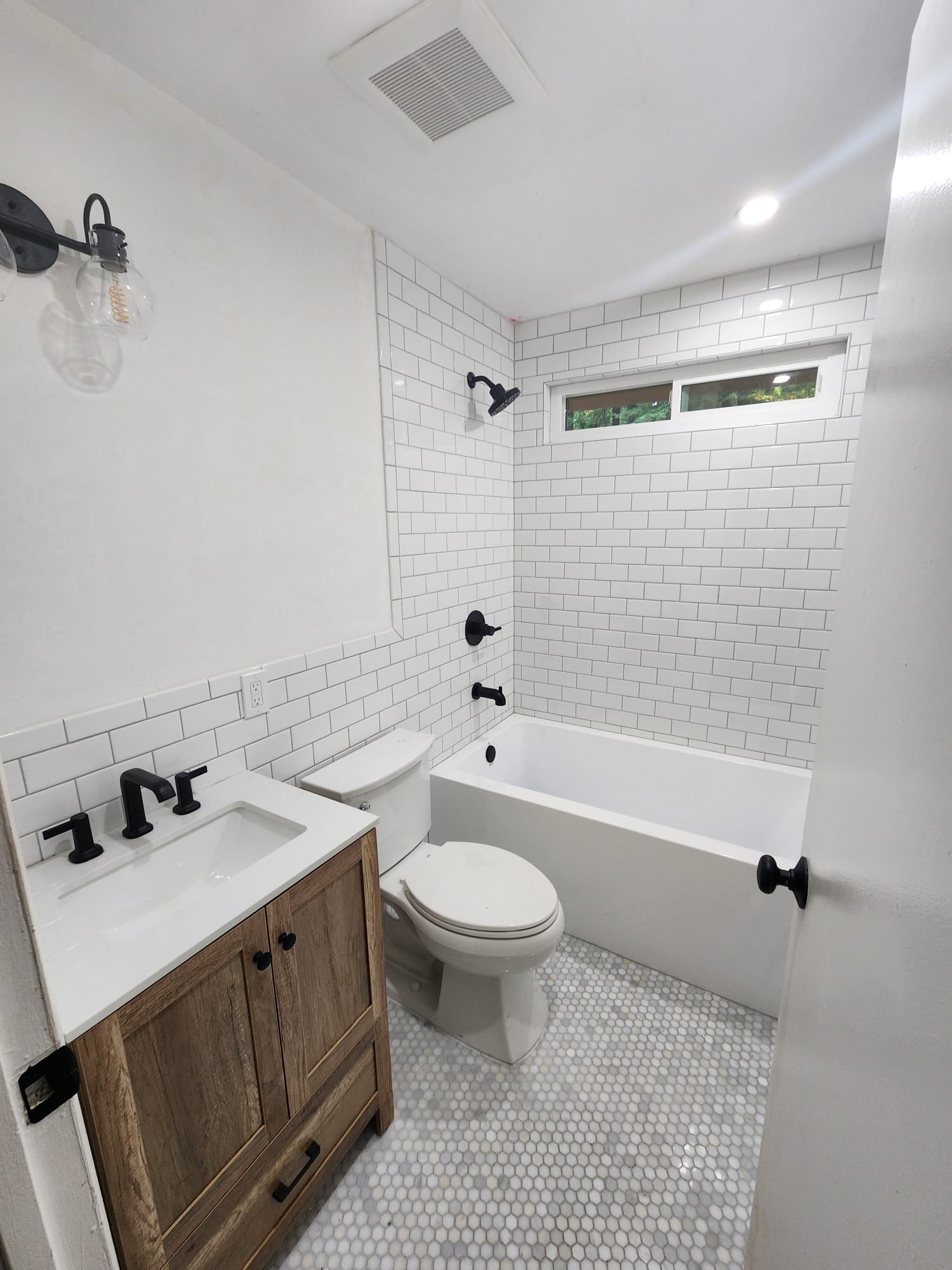 A small bathroom with a white-tiled bathtub, toilet, and sink. The sink has a wooden cabinet and black fixtures. The floor is covered in hexagonal tiles. One light fixture is mounted on the wall.
