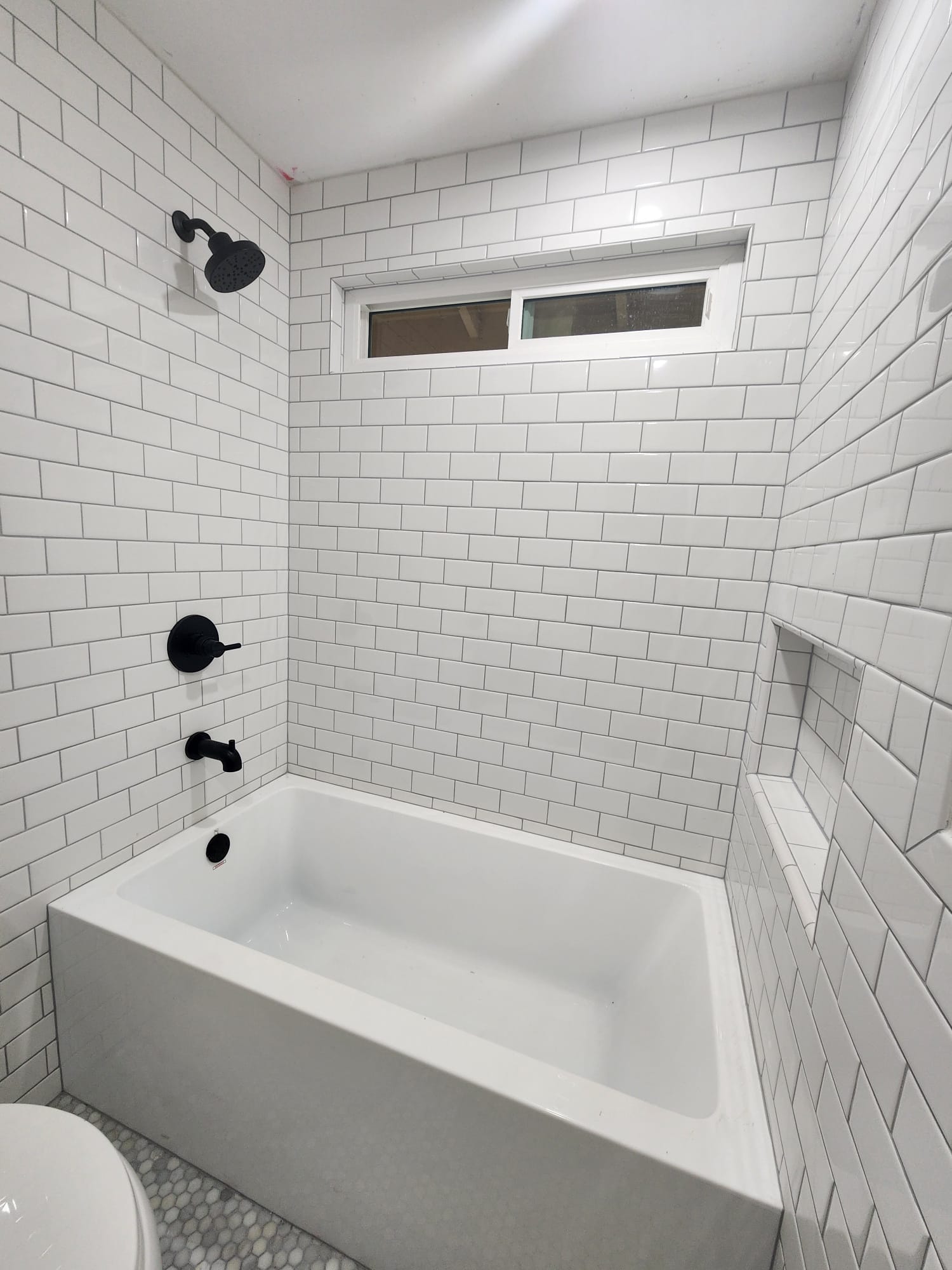 A modern bathroom features a white subway-tiled bath and shower area with a black showerhead and faucet, a rectangular window, and a built-in wall niche.