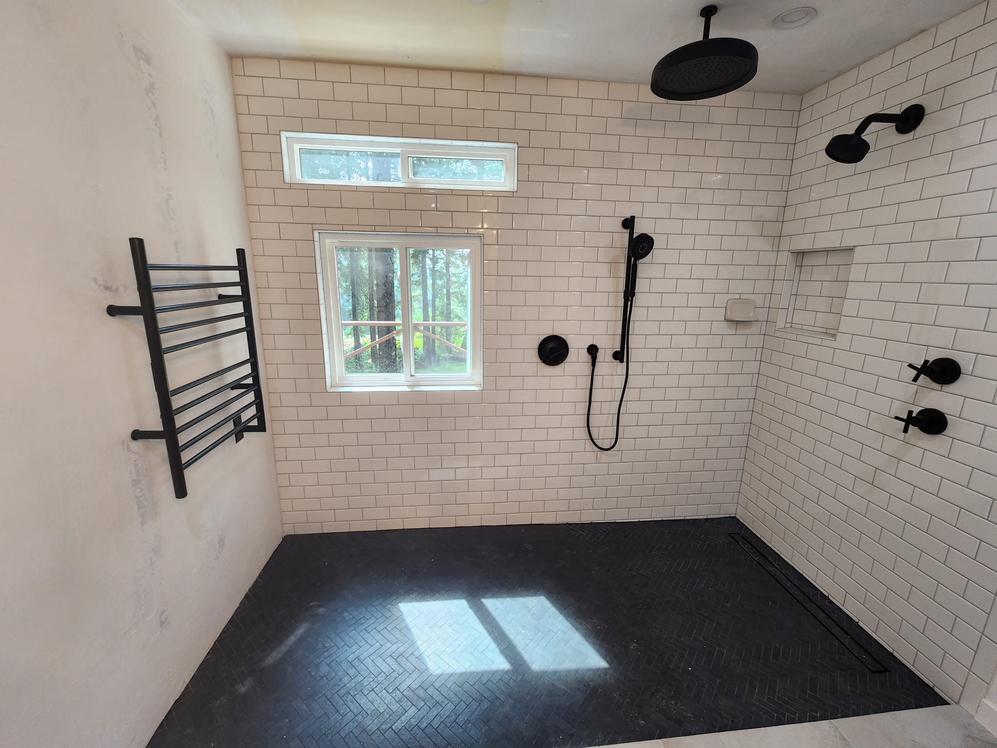 A bathroom with white subway tile walls, a window, black shower fixtures, a black towel rack, and a dark herringbone tile floor.