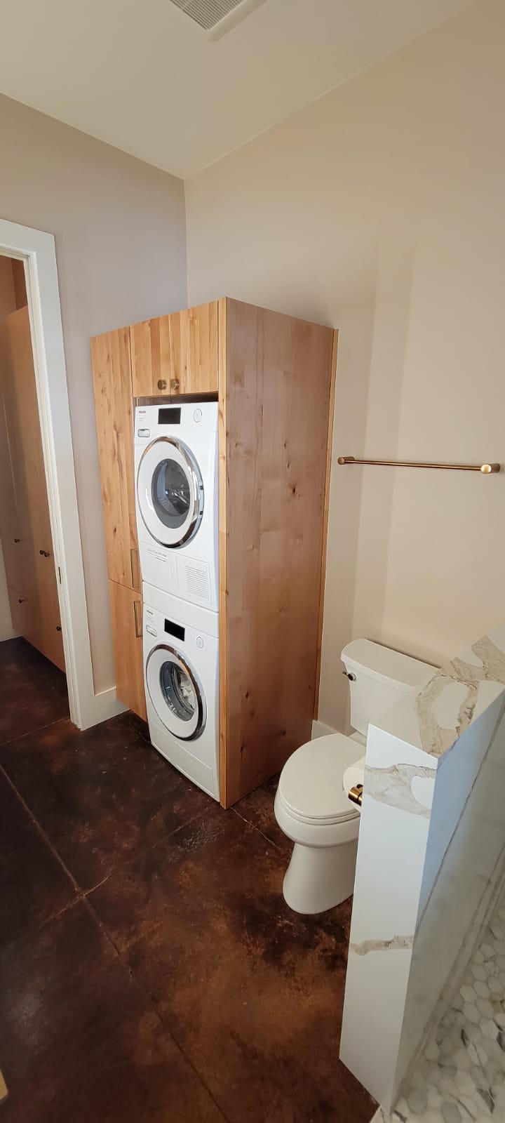 A bathroom with a stackable washer and dryer unit housed in wooden cabinetry next to a toilet and a marble-patterned half wall.