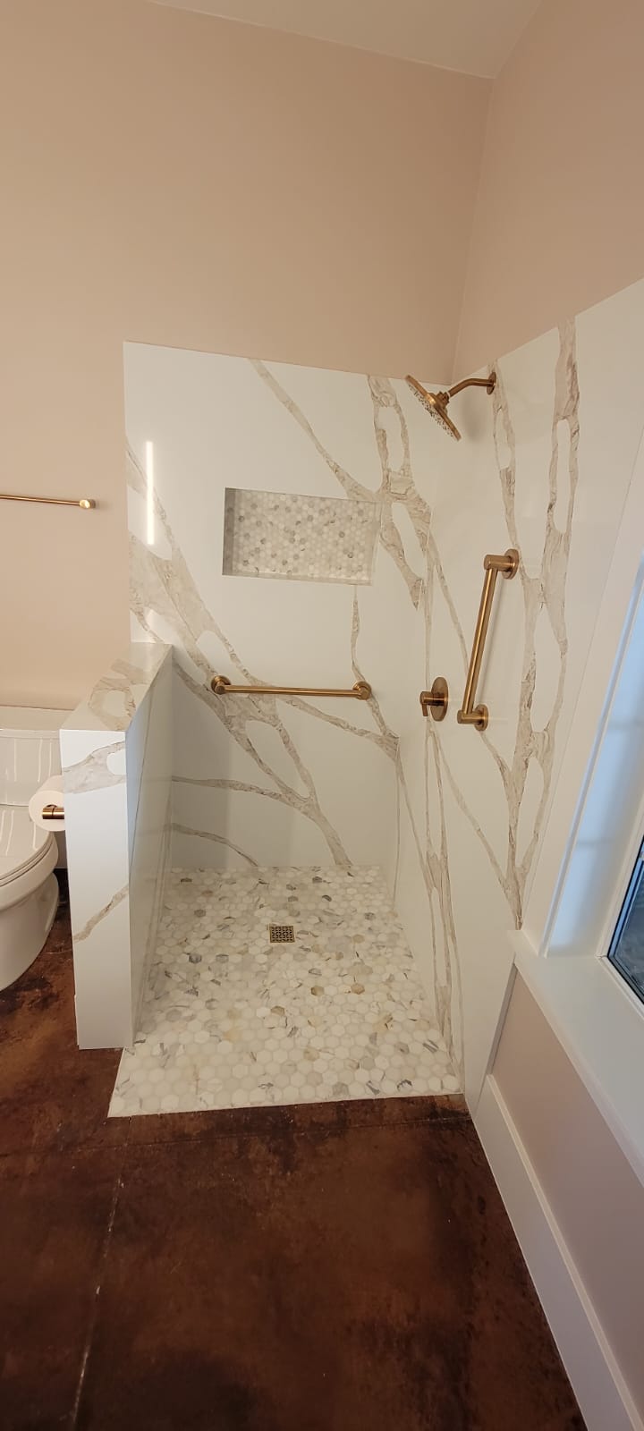 A modern walk-in shower with marble walls, built-in shelf, gold fixtures, and a white pebble floor. The adjacent area includes a wall-mounted bar and a toilet visible on the left.