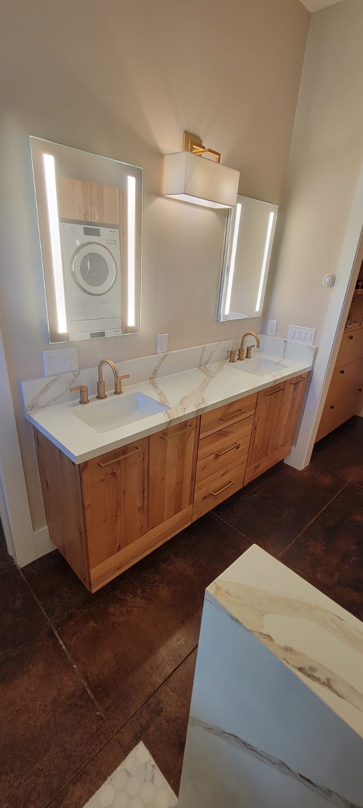 A bathroom with a double sink vanity, two mirrors with integrated lighting, and a washer dryer combo unit mounted on the wall.