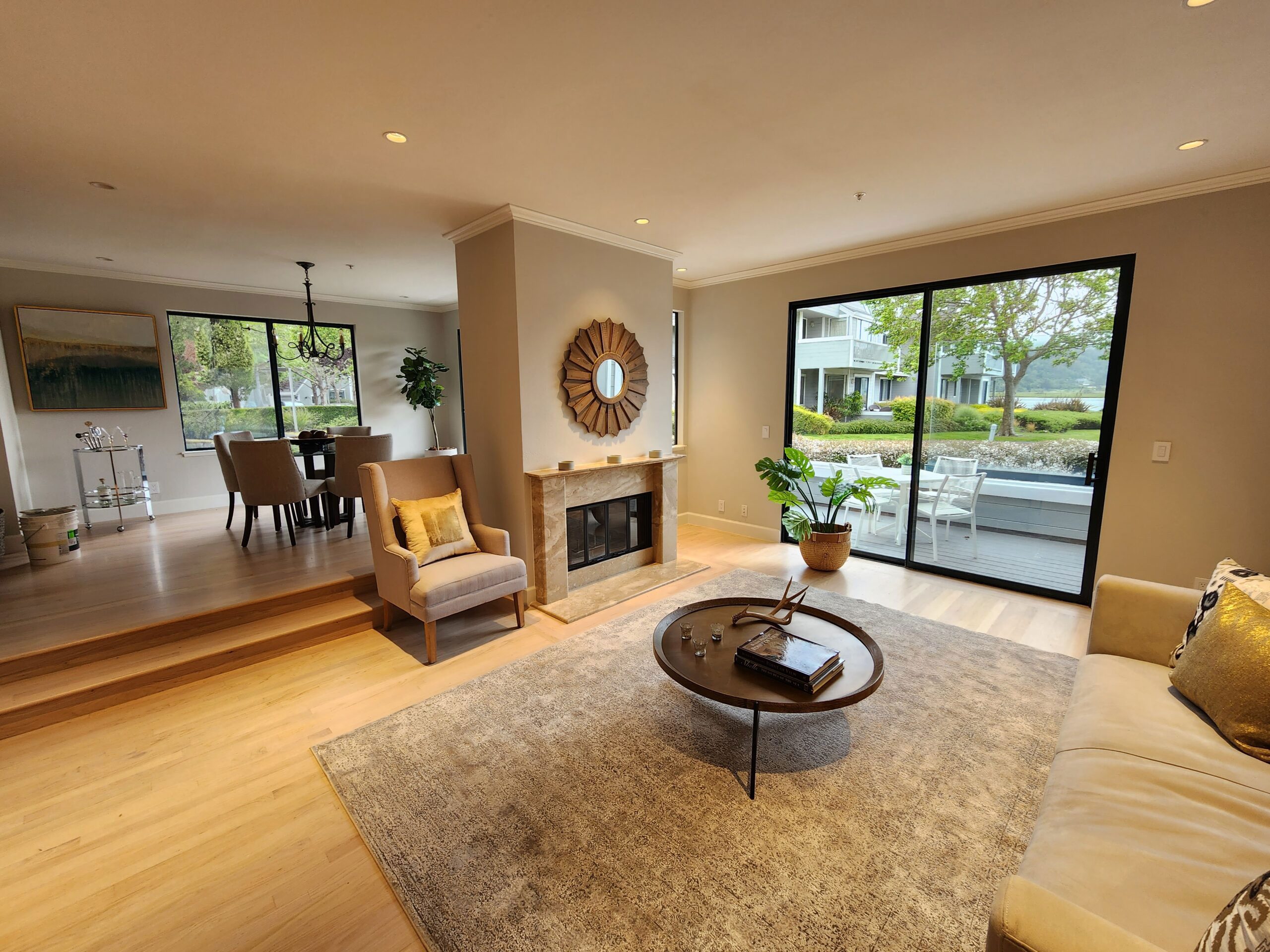 A modern living room with light wood flooring, a fireplace, a round mirror, and large sliding glass doors leading to an outdoor deck. The room is furnished with a beige sofa and armchairs.