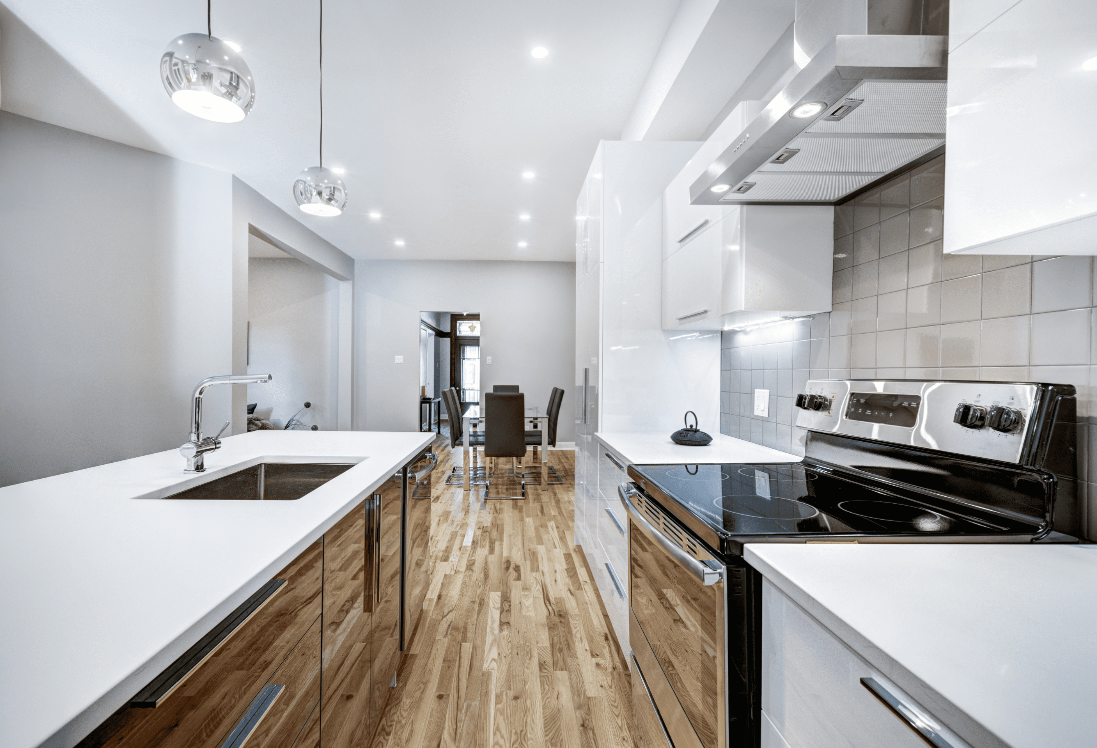 Modern kitchen with stainless steel appliances, polished wooden floors, white countertops, and white cabinetry. Two pendant lights hang over the island with a sink and faucet. Dining area in background.