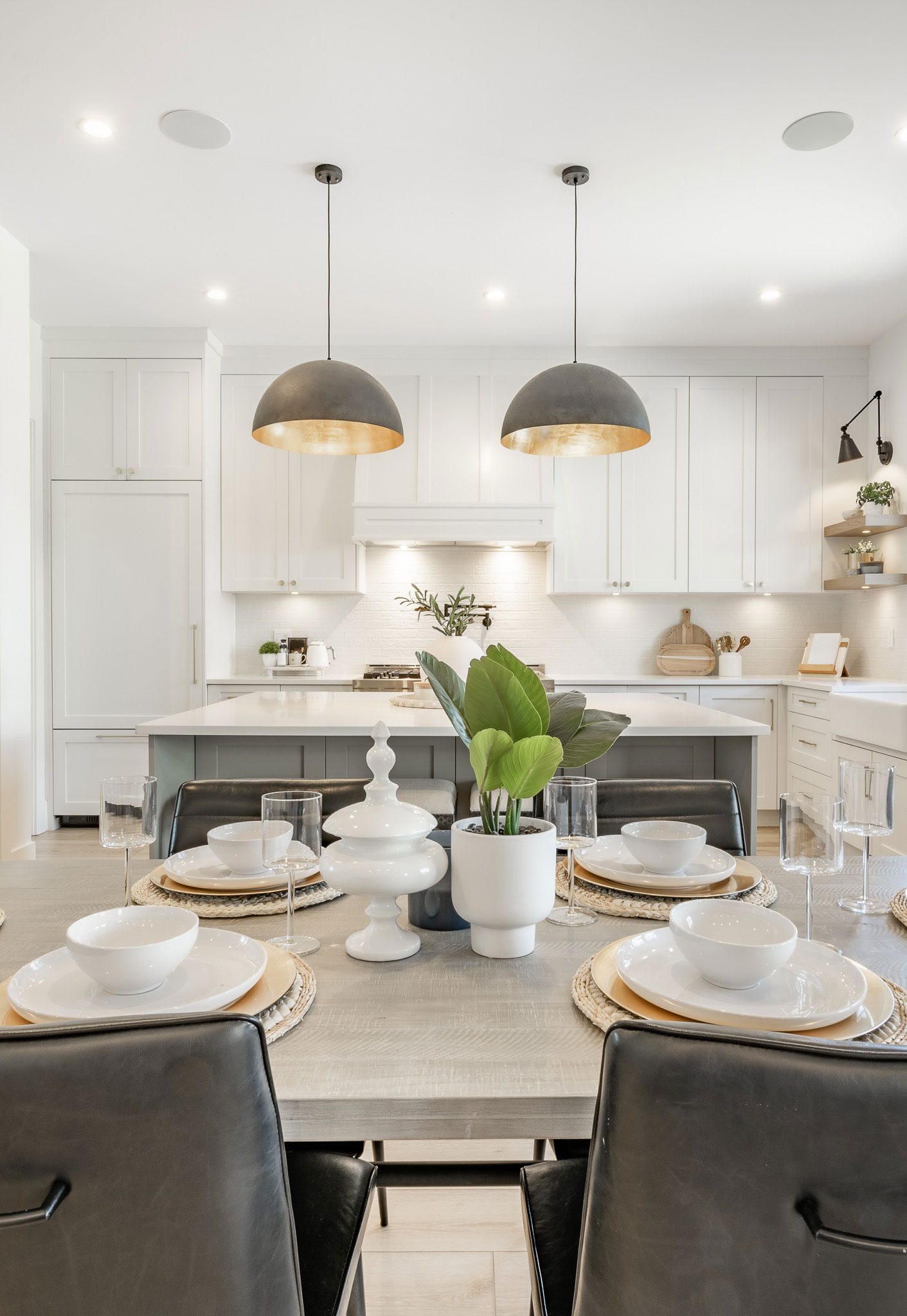Modern kitchen with white cabinets, a central island, pendant lights, and a dining table set with plates and bowls. A potted plant and decorative items are on the table.