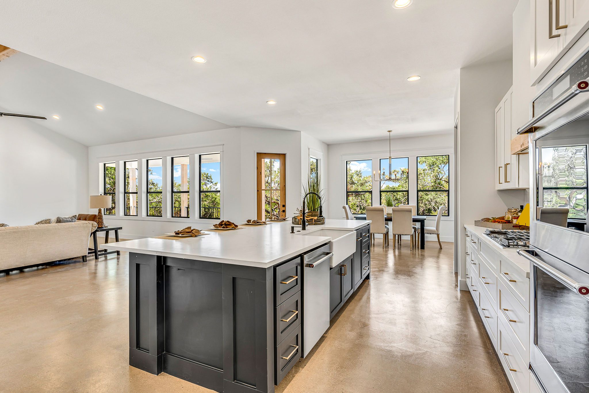 Modern kitchen and dining area featuring a large island with a sink, white cabinets, stainless steel appliances, and ample natural light from numerous windows and glass doors, with a living area adjacent.